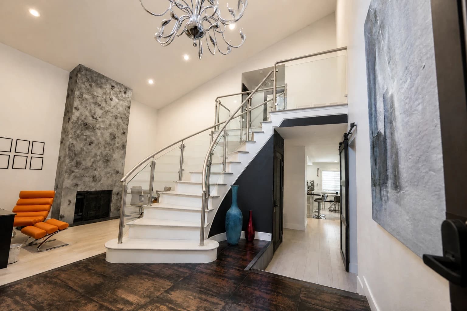 Bright modern interior with a curved white staircase, glass railings, stone fireplace, and orange lounge chair.