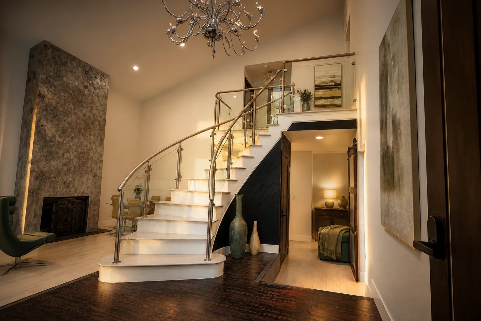 Residence foyer at dusk: a curved staircase with a brass-and-glass rail, a stone fireplace, and a chandelier overhead.