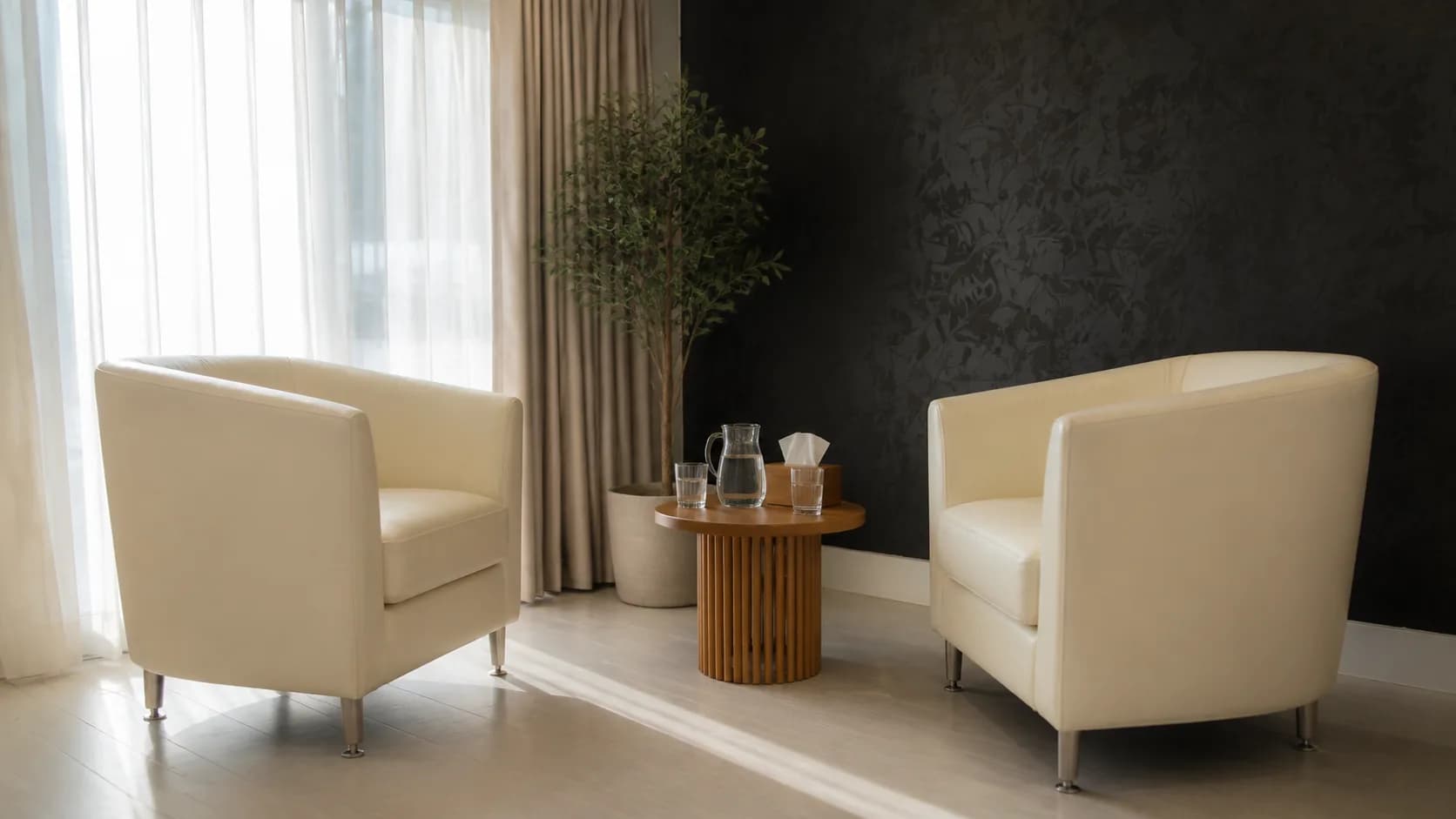 Two cream armchairs and a round side table beside a curtained window at Providence Detox.